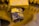 A close-up image of a metal bolt attached to a larger yellow structure, showing signs of rust and wear. The nut is partially threaded over the bolt. The surface is painted in yellow with visible patches of corrosion and chipping paint, surrounded by other metallic parts.