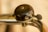 A close-up view of a metallic bicycle bell mounted on handlebars. The bell has a dark, weathered top with a visible screw in the center, indicating signs of use. The bell's lever is labeled with the word 'MINAR.’ The background is out of focus, creating a warm, slightly blurred effect.