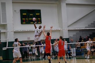 A volleyball game in progress with players in mid-air attempting a spike over the net. The player in white is jumping to hit the ball while opposing players in red jerseys attempt to block. The scene is set in an indoor sports arena with spectators in the background.