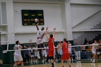 A volleyball game in progress with players in mid-air attempting a spike over the net. The player in white is jumping to hit the ball while opposing players in red jerseys attempt to block. The scene is set in an indoor sports arena with spectators in the background.