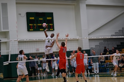 A volleyball game in progress with players in mid-air attempting a spike over the net. The player in white is jumping to hit the ball while opposing players in red jerseys attempt to block. The scene is set in an indoor sports arena with spectators in the background.