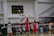 A volleyball game in progress with players in mid-air attempting a spike over the net. The player in white is jumping to hit the ball while opposing players in red jerseys attempt to block. The scene is set in an indoor sports arena with spectators in the background.