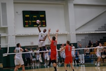 A volleyball game in progress with players in mid-air attempting a spike over the net. The player in white is jumping to hit the ball while opposing players in red jerseys attempt to block. The scene is set in an indoor sports arena with spectators in the background.