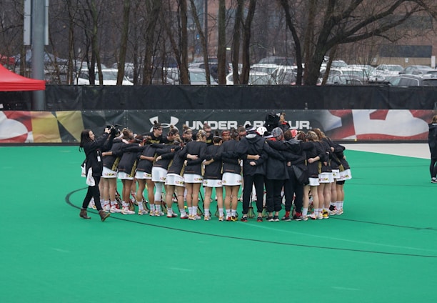A group of athletes is gathered in a huddle on a sports field, wearing matching uniforms and gear. The ground is green, and there is signage with a brand logo in the background. Trees and parked cars can be seen behind a fence surrounding the field. A person on the left side is capturing the moment with a camera.