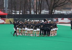 A group of athletes is gathered in a huddle on a sports field, wearing matching uniforms and gear. The ground is green, and there is signage with a brand logo in the background. Trees and parked cars can be seen behind a fence surrounding the field. A person on the left side is capturing the moment with a camera.