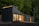 A modern, dark-colored, modular home situated in a lush forest environment. Large windows allow warm interior light to spill out, contrasting with the natural greenery surrounding the structure. The building has a sleek, geometric design, with a sloping roof and a wooden deck that extends from its side.