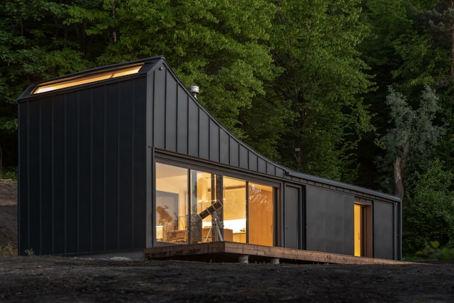 A modern, dark-colored, modular home situated in a lush forest environment. Large windows allow warm interior light to spill out, contrasting with the natural greenery surrounding the structure. The building has a sleek, geometric design, with a sloping roof and a wooden deck that extends from its side.