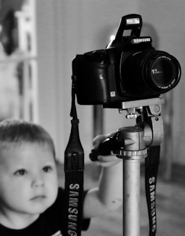 A child is looking at a DSLR camera mounted on a tripod. The camera has a strap with the brand name Samsung visible. The image is in black and white, emphasizing the focus on the camera and the child's curious expression.