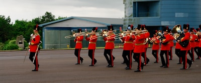 A marching band dressed in red military-style uniforms, including gold embellishments and black hats, is performing on a large paved area. The musicians are playing brass instruments such as trombones and trumpets. A building with a metallic exterior and large windows is visible in the background, along with some greenery and overcast skies.