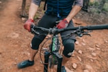 A cyclist wearing gloves and knee pads is gripping the handlebars of a mountain bike. The bike is on a rugged dirt trail, and the cyclist's lower half is visible.