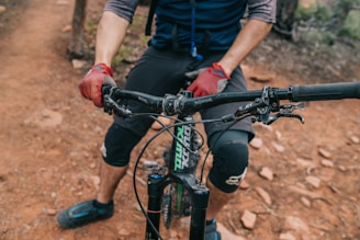 A cyclist wearing gloves and knee pads is gripping the handlebars of a mountain bike. The bike is on a rugged dirt trail, and the cyclist's lower half is visible.