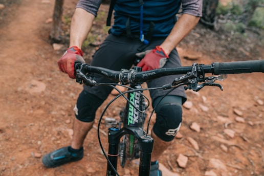 A cyclist wearing gloves and knee pads is gripping the handlebars of a mountain bike. The bike is on a rugged dirt trail, and the cyclist's lower half is visible.