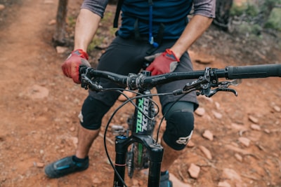 A cyclist wearing gloves and knee pads is gripping the handlebars of a mountain bike. The bike is on a rugged dirt trail, and the cyclist's lower half is visible.