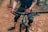A cyclist wearing gloves and knee pads is gripping the handlebars of a mountain bike. The bike is on a rugged dirt trail, and the cyclist's lower half is visible.
