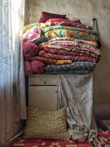 A stack of colorful, patterned quilts and pillows is piled on top of a vintage, chipped dresser in a room with textured, aging walls. The ornate, lace curtains allow soft light to filter through, casting gentle shadows. A patterned pillow rests on the floor against the dresser, and a floral-patterned rug adds warmth to the space.