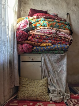 A stack of colorful, patterned quilts and pillows is piled on top of a vintage, chipped dresser in a room with textured, aging walls. The ornate, lace curtains allow soft light to filter through, casting gentle shadows. A patterned pillow rests on the floor against the dresser, and a floral-patterned rug adds warmth to the space.