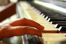 A hand gently rests on the keys of a piano, focusing on the interaction between the fingers and the ivory keys. The image captures the texture of the skin and the grain of the wooden piano.