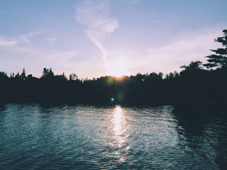 A serene lake reflecting the soft glow of a setting sun, surrounded by whispering trees.