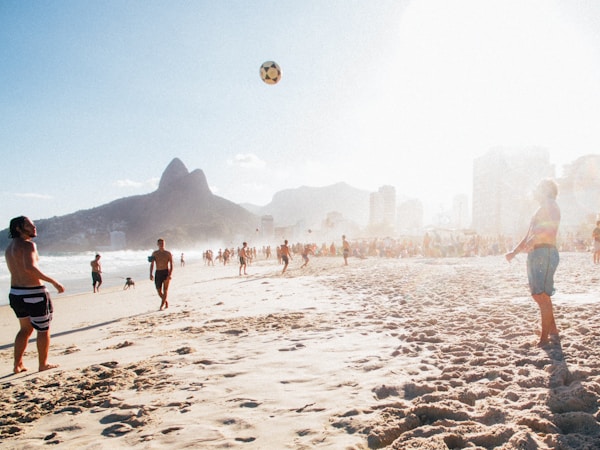 A sunlit beach scene with people playing with a soccer ball. The shoreline is bustling with activity, and the backdrop features mountains and tall buildings. The bright sunlight creates a warm and inviting atmosphere.