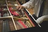 A person in a white shirt and a tie is using a tuning wrench to adjust the strings inside a grand piano. The interior of the piano, including its strings and red felt lining, is visible. The person's other hand is gloved and resting on the edge of the piano.