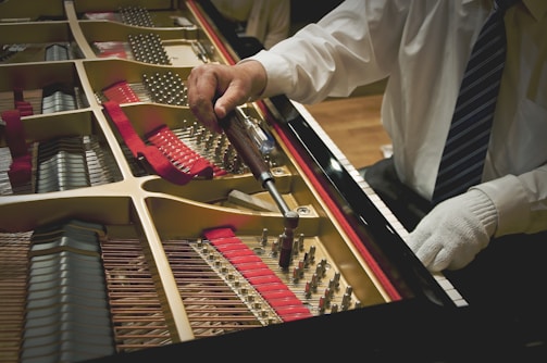 A person in a white shirt and a tie is using a tuning wrench to adjust the strings inside a grand piano. The interior of the piano, including its strings and red felt lining, is visible. The person's other hand is gloved and resting on the edge of the piano.
