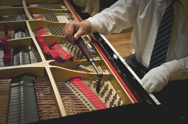 A person in a white shirt and a tie is using a tuning wrench to adjust the strings inside a grand piano. The interior of the piano, including its strings and red felt lining, is visible. The person's other hand is gloved and resting on the edge of the piano.