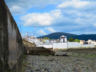 A seaside town's coastal wall and rocky shore are in the foreground, with a row of colorful buildings lining the waterfront. A green hill and forested areas are visible in the distance, under a blue sky filled with fluffy white clouds.