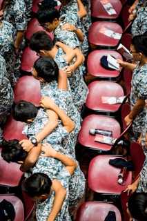 A group of young individuals wearing camouflage uniforms are huddled together in a friendly, supportive manner. They are in a large room with red chairs arranged in rows. Some of the chairs have pamphlets and water bottles placed on them.