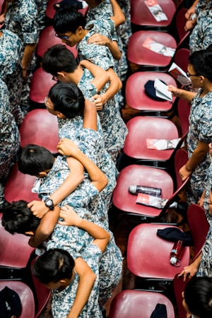A group of young individuals wearing camouflage uniforms are huddled together in a friendly, supportive manner. They are in a large room with red chairs arranged in rows. Some of the chairs have pamphlets and water bottles placed on them.