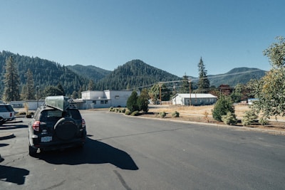 A parking lot with several vehicles, including a black SUV with a canoe mounted on top. In the background, there are industrial buildings and residential houses surrounded by trees. Beyond, forested hills under a clear blue sky provide a scenic backdrop.