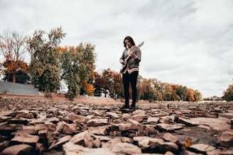 A person stands on a rocky surface with an electric guitar in their hands. The background features trees, some with autumn-colored leaves, under a cloudy sky.