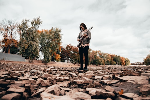 A person stands on a rocky surface with an electric guitar in their hands. The background features trees, some with autumn-colored leaves, under a cloudy sky.