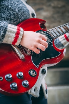 A person is playing a red electric guitar, focusing on the hand positioned over the guitar strings. The guitar has a glossy finish, visible knobs, and a switch labeled 'rhythm' and 'treble'. The person is wearing a cozy, striped sweater.
