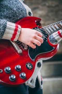A person is playing a red electric guitar, focusing on the hand positioned over the guitar strings. The guitar has a glossy finish, visible knobs, and a switch labeled 'rhythm' and 'treble'. The person is wearing a cozy, striped sweater.