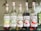 Five bottles of Monin syrup are lined up on a shelf. Each bottle has a different label indicating its flavor: Guava, Mojito Mint, Litchi, Fraise, and Watermelon. The labels display colorful illustrations of the respective fruits or ingredients. The bottles are made of clear glass and are topped with white and red caps. The background is slightly blurred, suggesting an indoor setting with elements of greenery.