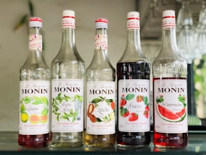 Five bottles of Monin syrup are lined up on a shelf. Each bottle has a different label indicating its flavor: Guava, Mojito Mint, Litchi, Fraise, and Watermelon. The labels display colorful illustrations of the respective fruits or ingredients. The bottles are made of clear glass and are topped with white and red caps. The background is slightly blurred, suggesting an indoor setting with elements of greenery.