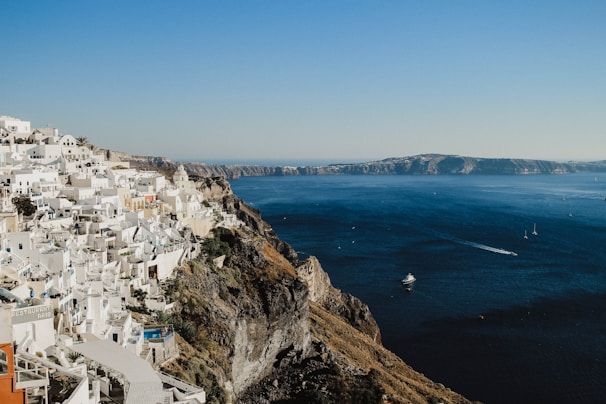 White buildings with blue domes cascade down a steep cliffside, overlooking a vast, serene blue sea. The coastline stretches out in the distance under a clear sky, with a few small boats dotting the water.