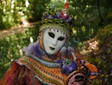 An individual wears an ornate carnival mask and costume adorned with intricate beadwork and vibrant colors, standing amidst a lush, green natural backdrop. The costume features bright orange, purple, and green hues, embellished with sequins and feathers. The mask has a blank expression with dark, captivating eye makeup, contributing to the mysterious and theatrical appearance.