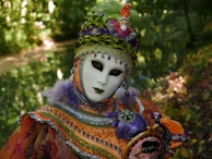 An individual wears an ornate carnival mask and costume adorned with intricate beadwork and vibrant colors, standing amidst a lush, green natural backdrop. The costume features bright orange, purple, and green hues, embellished with sequins and feathers. The mask has a blank expression with dark, captivating eye makeup, contributing to the mysterious and theatrical appearance.