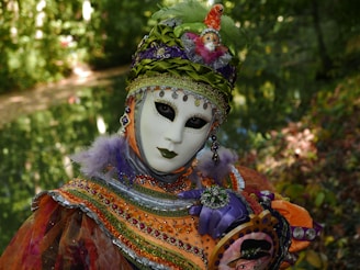 An individual wears an ornate carnival mask and costume adorned with intricate beadwork and vibrant colors, standing amidst a lush, green natural backdrop. The costume features bright orange, purple, and green hues, embellished with sequins and feathers. The mask has a blank expression with dark, captivating eye makeup, contributing to the mysterious and theatrical appearance.