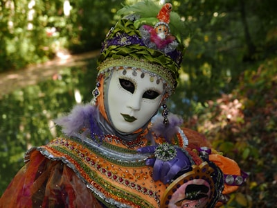 An individual wears an ornate carnival mask and costume adorned with intricate beadwork and vibrant colors, standing amidst a lush, green natural backdrop. The costume features bright orange, purple, and green hues, embellished with sequins and feathers. The mask has a blank expression with dark, captivating eye makeup, contributing to the mysterious and theatrical appearance.