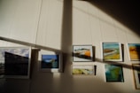 A series of framed paintings are displayed on a wall. Sunlight creates contrasting shadows across the wall, highlighting the artwork. The paintings feature various landscapes and abstract scenes with a mix of colors.