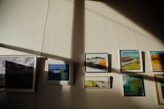 A series of framed paintings are displayed on a wall. Sunlight creates contrasting shadows across the wall, highlighting the artwork. The paintings feature various landscapes and abstract scenes with a mix of colors.