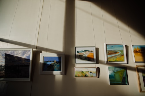 A series of framed paintings are displayed on a wall. Sunlight creates contrasting shadows across the wall, highlighting the artwork. The paintings feature various landscapes and abstract scenes with a mix of colors.
