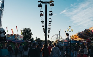 A lively carnival scene with a central view of a sky ride featuring colorful gondolas. Crowds of people walk along the pathway, surrounded by various amusement park attractions. Brightly lit stalls and rides line both sides of the walkway, including a ferris wheel in the background. A large stuffed panda bear hangs as a prize at one stall, and banners or flags add to the festive atmosphere.