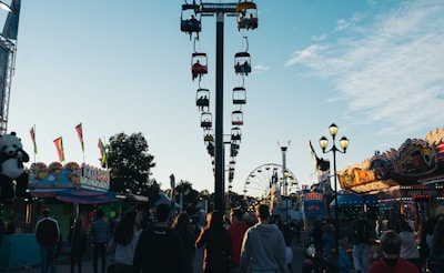 A lively carnival scene with a central view of a sky ride featuring colorful gondolas. Crowds of people walk along the pathway, surrounded by various amusement park attractions. Brightly lit stalls and rides line both sides of the walkway, including a ferris wheel in the background. A large stuffed panda bear hangs as a prize at one stall, and banners or flags add to the festive atmosphere.