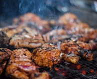 Juicy, well-seasoned pieces of grilled meat sizzle on a metal grill. The meat appears to be thoroughly cooked with a nice char and grill marks. Smoky wisps rise gently, adding an enticing aroma to the scene. The glowing red coals beneath suggest the meat is being cooked to perfection over an open flame.