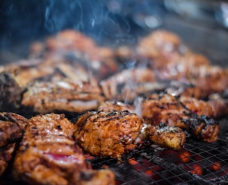 Juicy, well-seasoned pieces of grilled meat sizzle on a metal grill. The meat appears to be thoroughly cooked with a nice char and grill marks. Smoky wisps rise gently, adding an enticing aroma to the scene. The glowing red coals beneath suggest the meat is being cooked to perfection over an open flame.