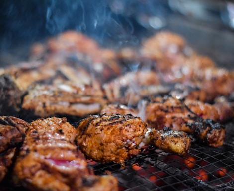 Juicy, well-seasoned pieces of grilled meat sizzle on a metal grill. The meat appears to be thoroughly cooked with a nice char and grill marks. Smoky wisps rise gently, adding an enticing aroma to the scene. The glowing red coals beneath suggest the meat is being cooked to perfection over an open flame.