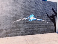 A brick wall painted dark gray with a vibrant blue and white spray paint art in the center. The artwork resembles an electric pulse or abstract energy flowing horizontally across the wall. To the right, a shadow of a hand creates a playful or dramatic effect.